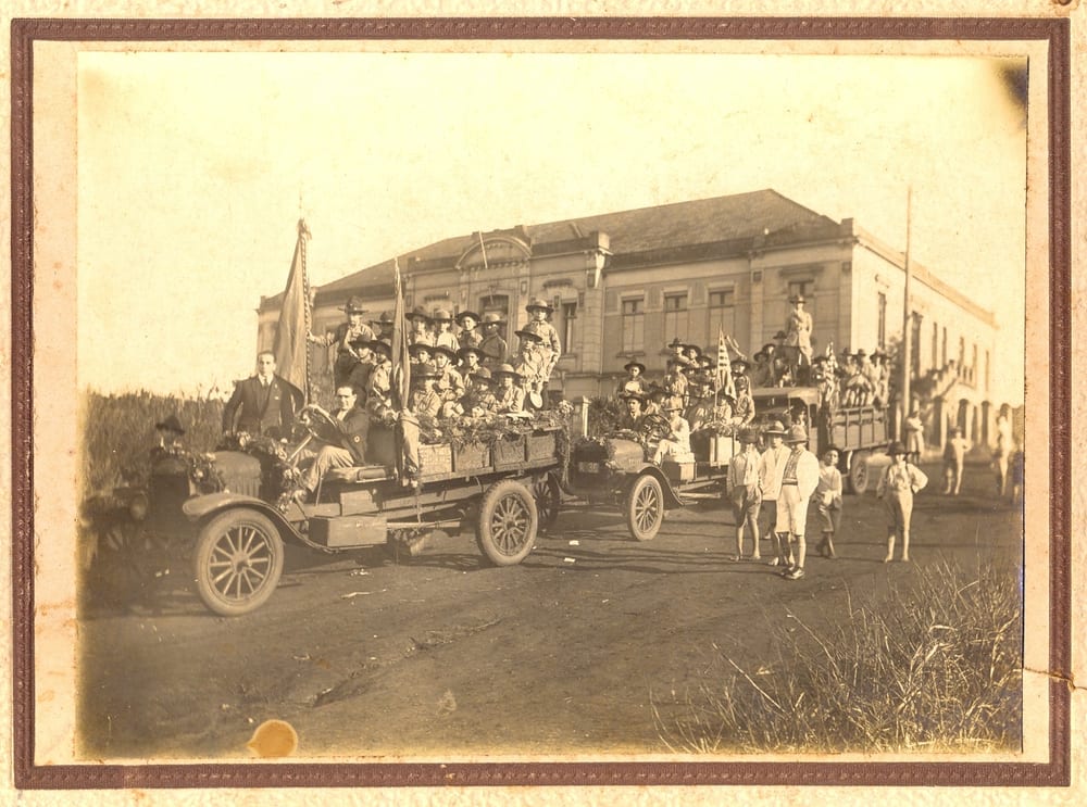 Caminhões enfeitados, cheios de crianças, diante do prédio do Grupo Escolar em dia de festa.